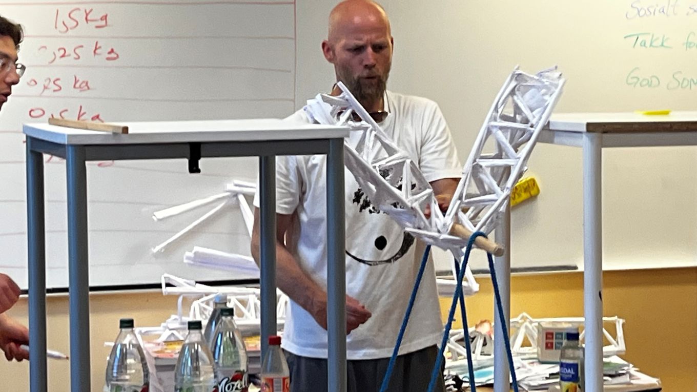 En lærer står ved en modell av ei bru bygd i papir. Brua er belastet med vekt, og her bryter den sammen.