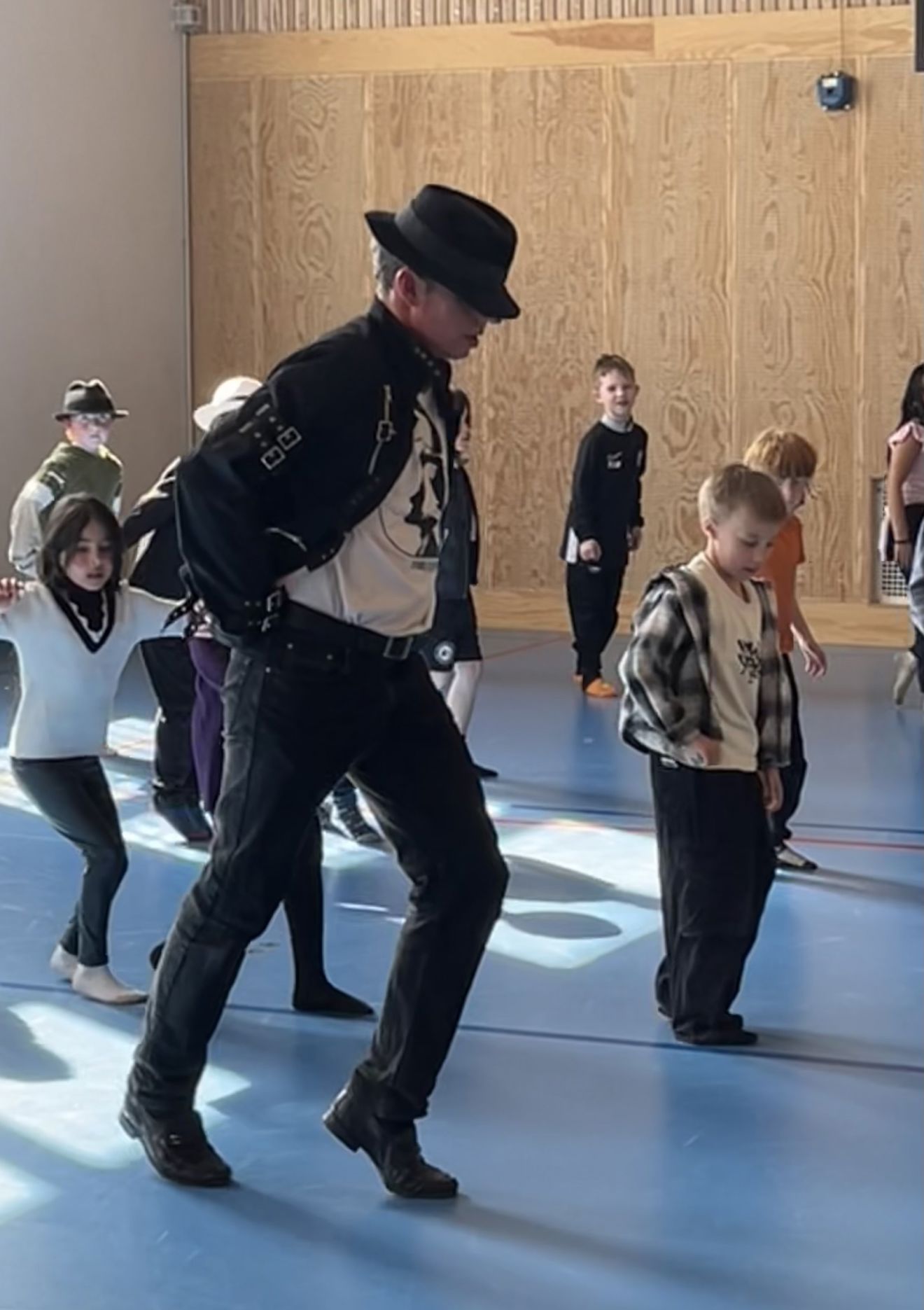En person med svart hatt og svart jakke danser med ei gruppe barn i en gymsal.