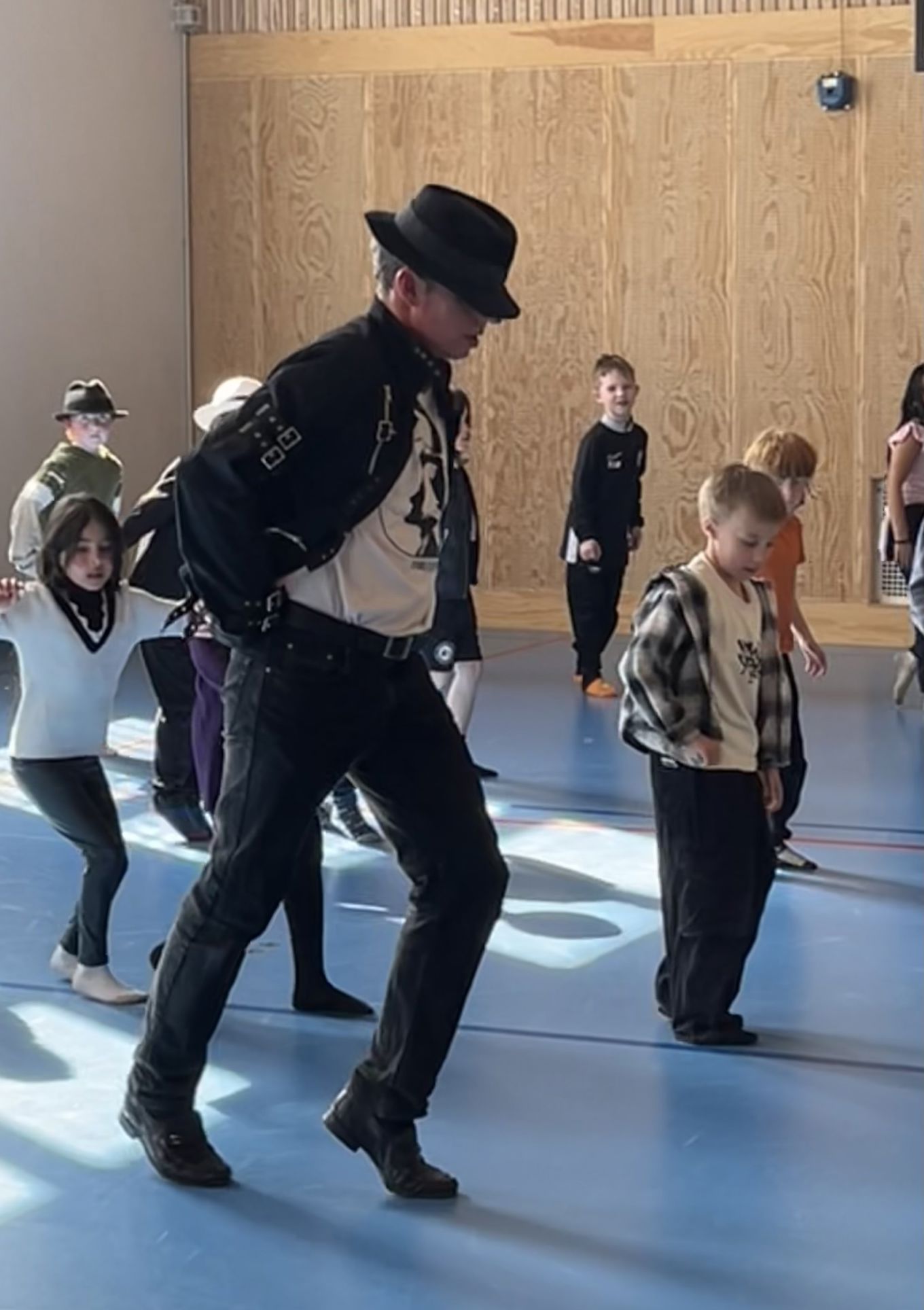 En person med svart hatt og svart jakke danser med ei gruppe barn i en gymsal.