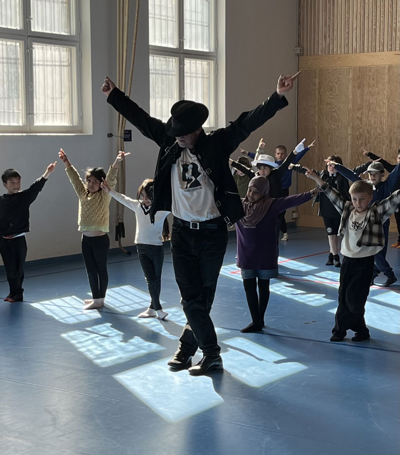 En instruktør og ei gruppe med elever danser. Sola skinner inn gjennom vinduet og på gulvet der de danser.