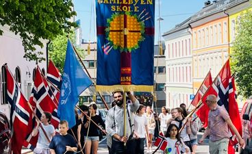 Elever går i øvetog i Arups gate før 17. mai. Vi ser skolens blå fane og flaggborgen.
