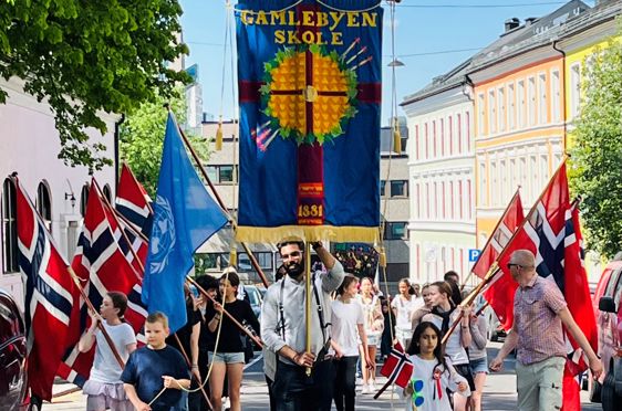 Elever går i øvetog i Arups gate før 17. mai. Vi ser skolens blå fane og flaggborgen.