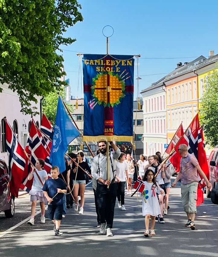 Elever går i øvetog i Arups gate før 17. mai. Vi ser skolens blå fane og flaggborgen.