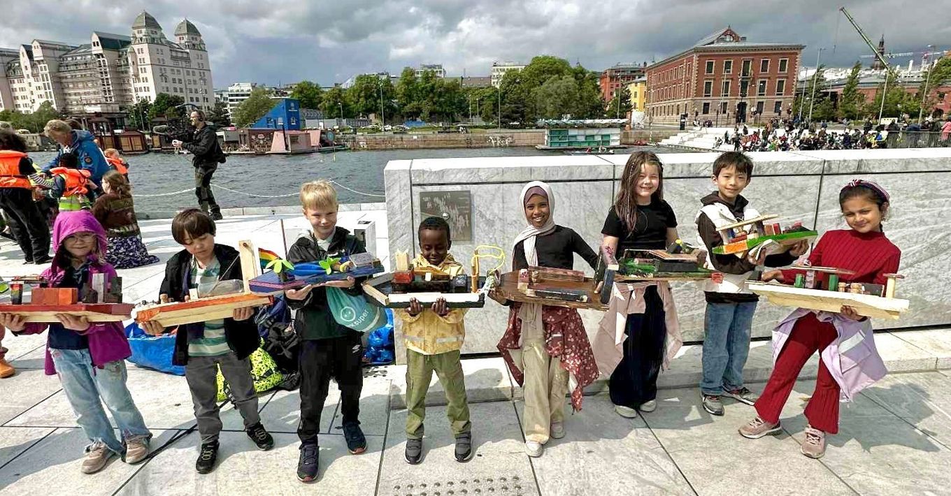 Ei gruppe elever som holder modellbåter. De står ved Operaen i Oslo med fjorden i bakgrunnen.