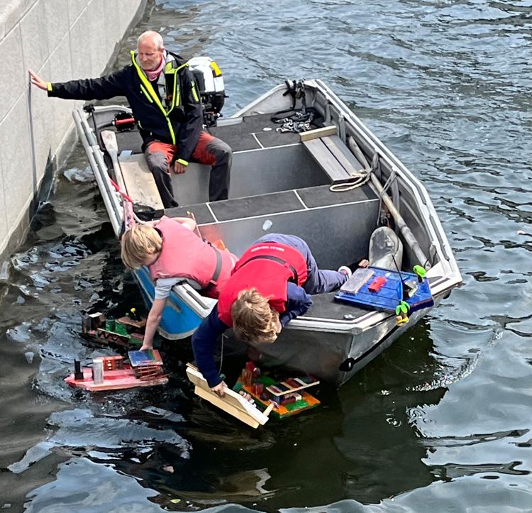 En lærer og to elever om bord i en åpen båt. De fisker modellbåter opp av vannet.