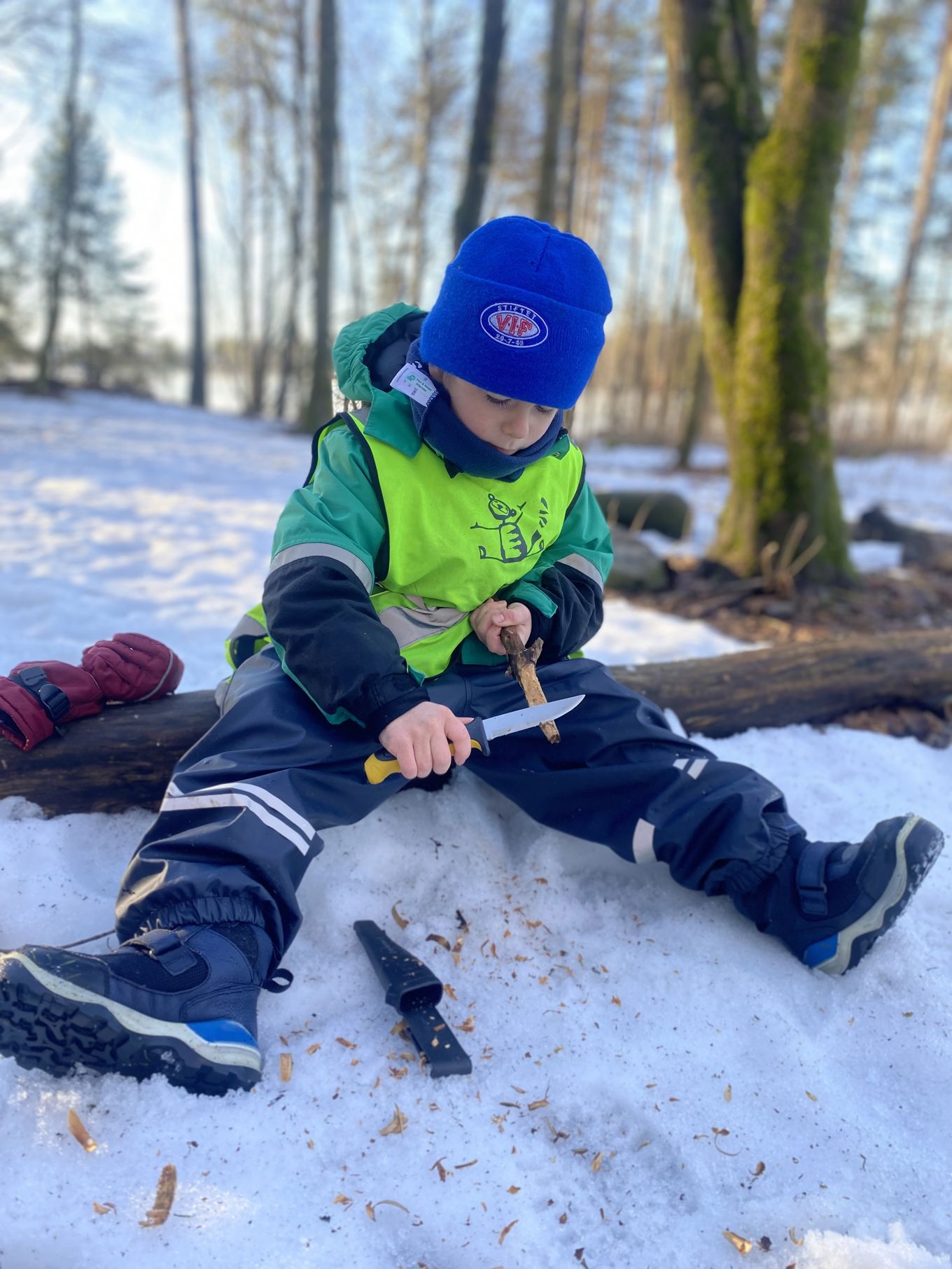 En gutt sitter på en stokk og spikker på en pinne. Det er vinter.