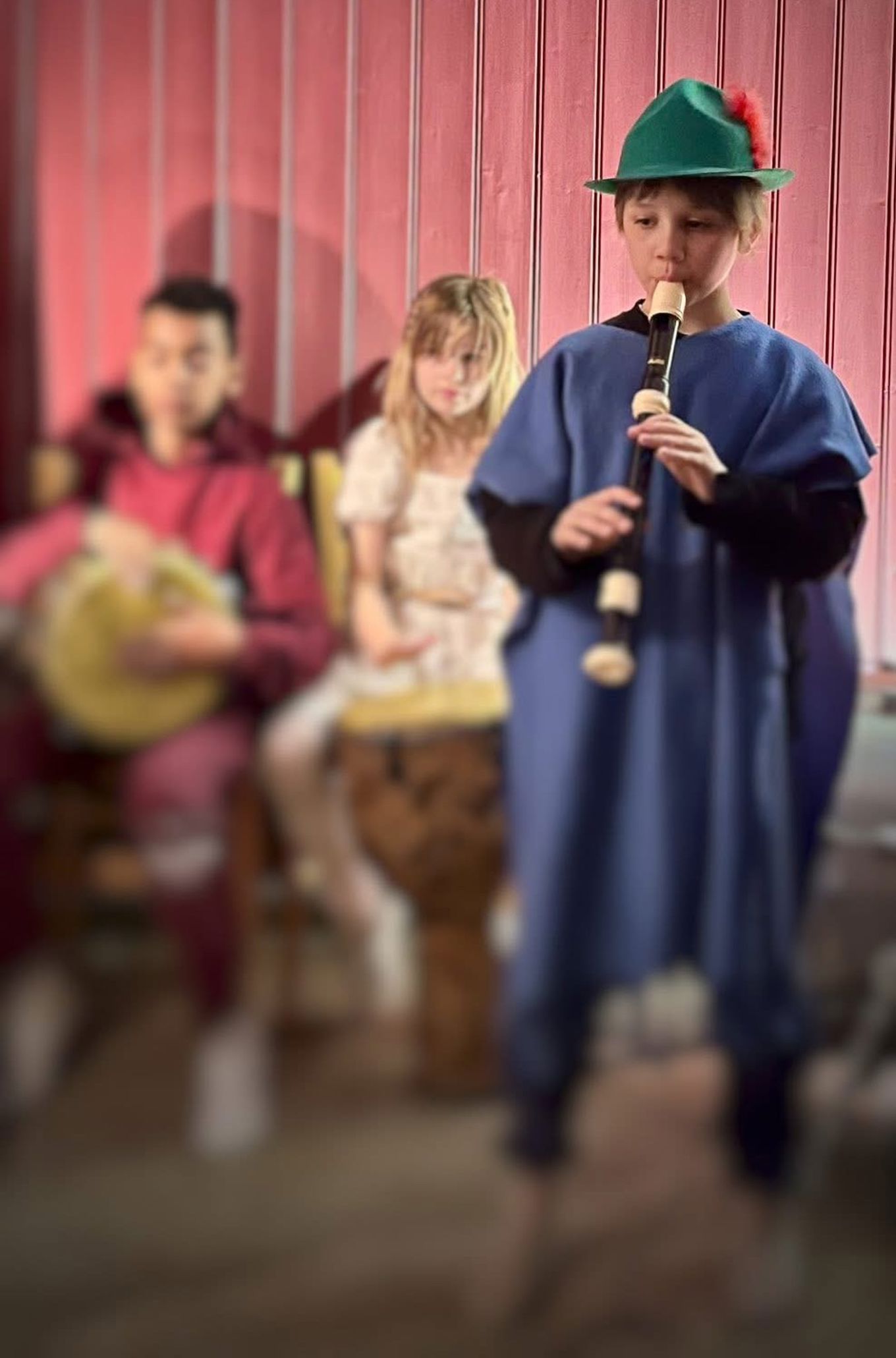 En elev med grønn hatt og blå kjortel står og spiller fløyte. To andre sitter i bakgrunnen og spiller trommer.