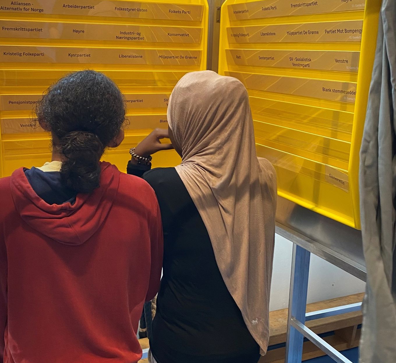 To elever står inne i et stemmeavlukke og ser på hyllene til stemmesedlene.