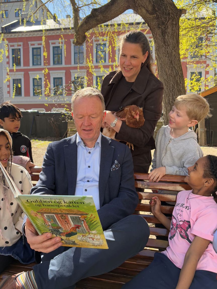 Rektor leser ei bok for ei gruppe barn ute i skolehagen mens sola skinner.