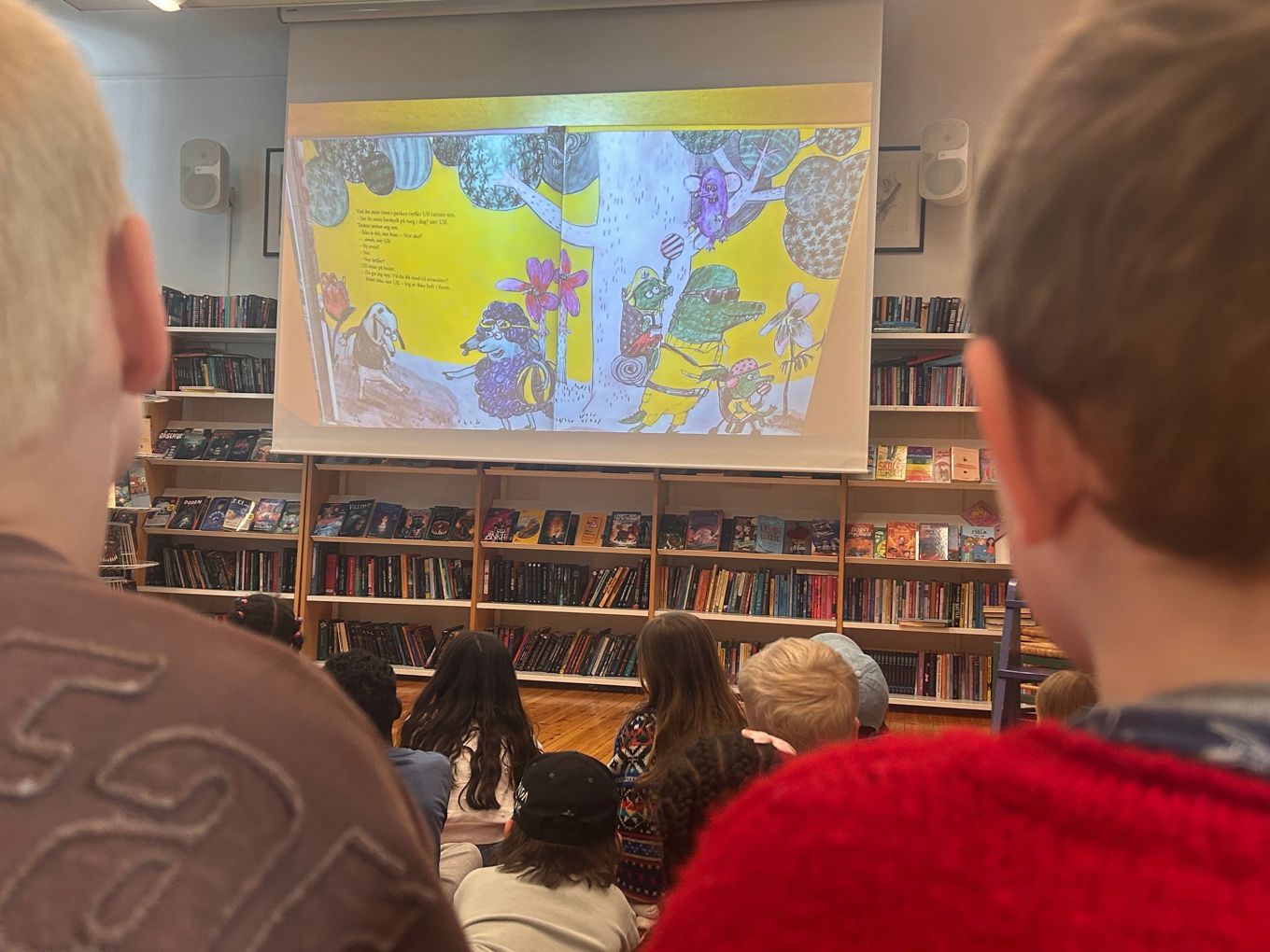 Førsteklassinger sitter på skolebiblioteket og følger med på høytlesing fra ei bok. Boka vises på en skjerm.